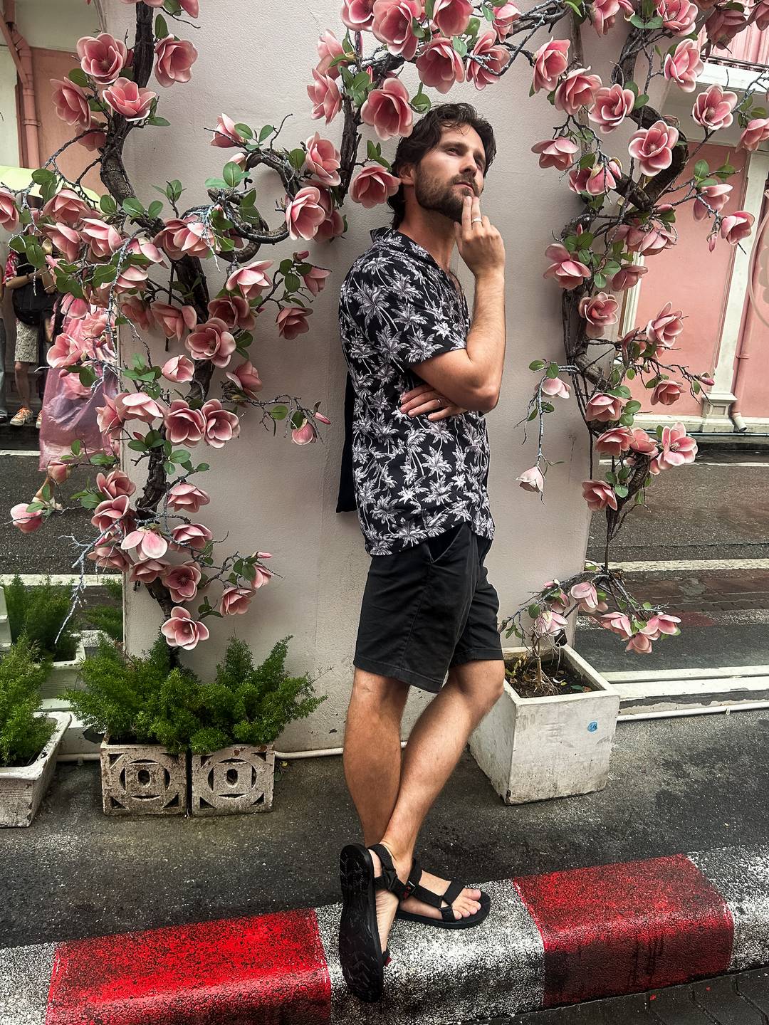 A man in a black and white shirt jokingly doing a pensive pose infront of a wall covered in pink flowers