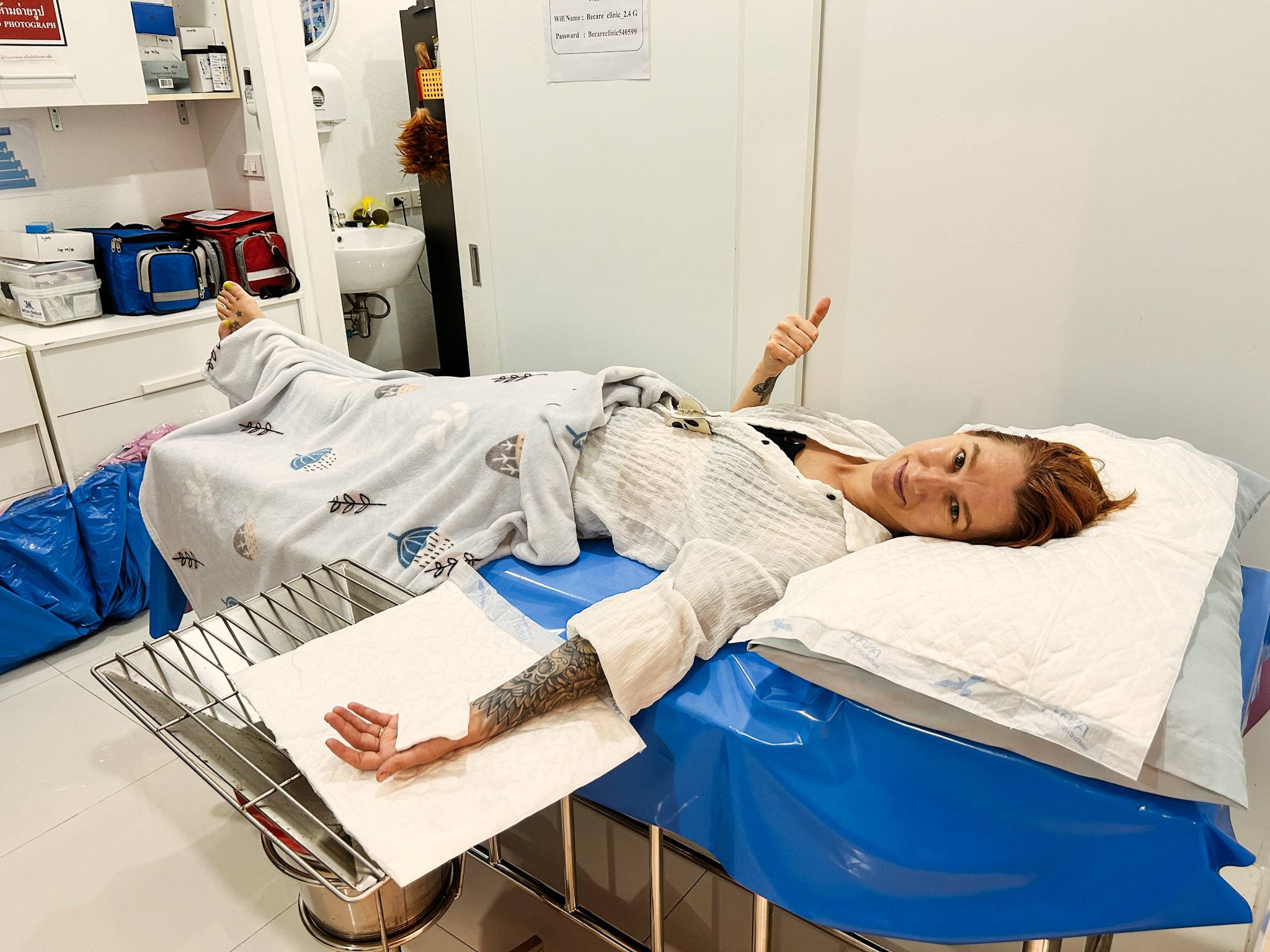a woman lying on a hospital bed, looking at the camera with her thump up