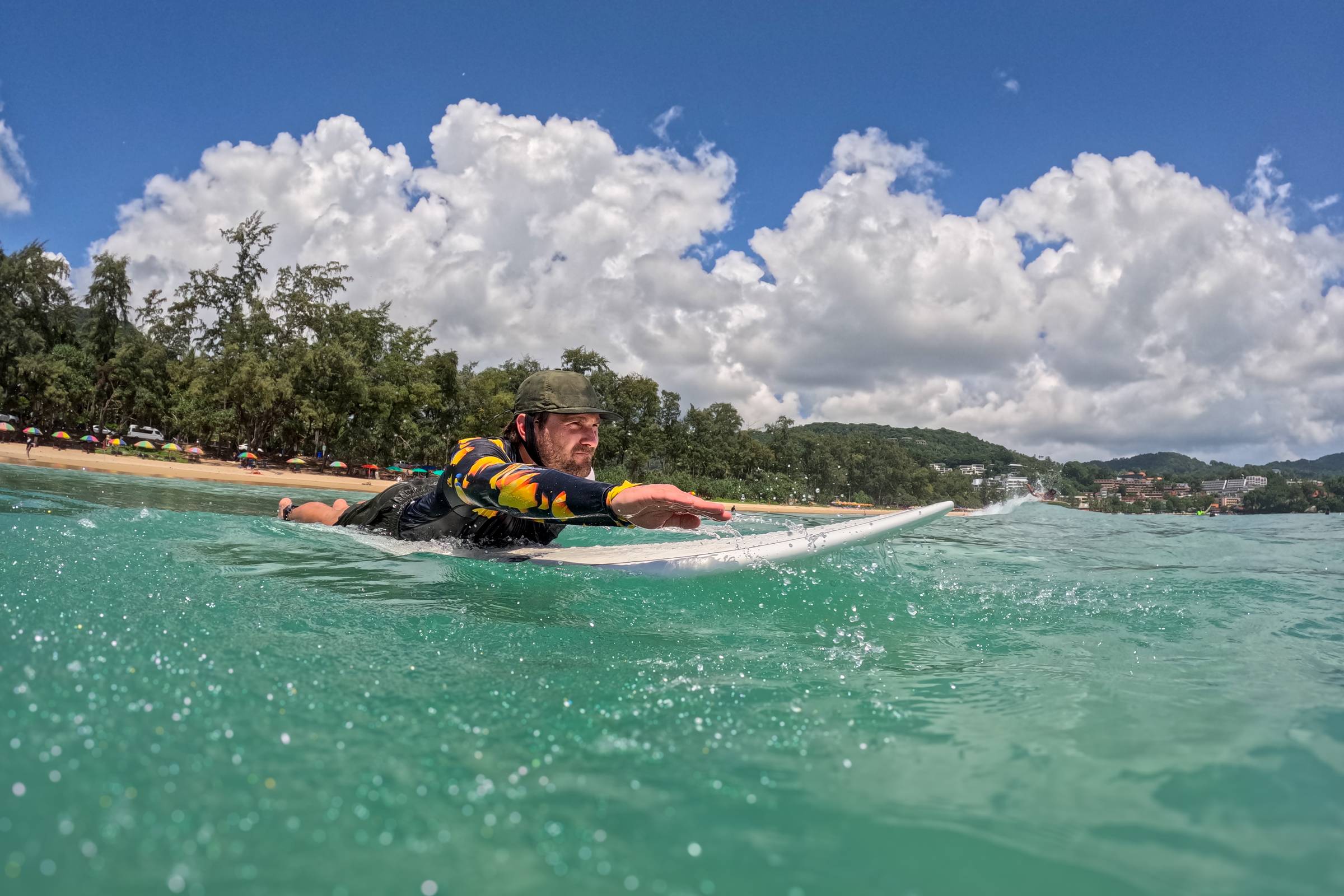 Man paddling on his surfboard in light blue waters with a cloudy blue sky above.