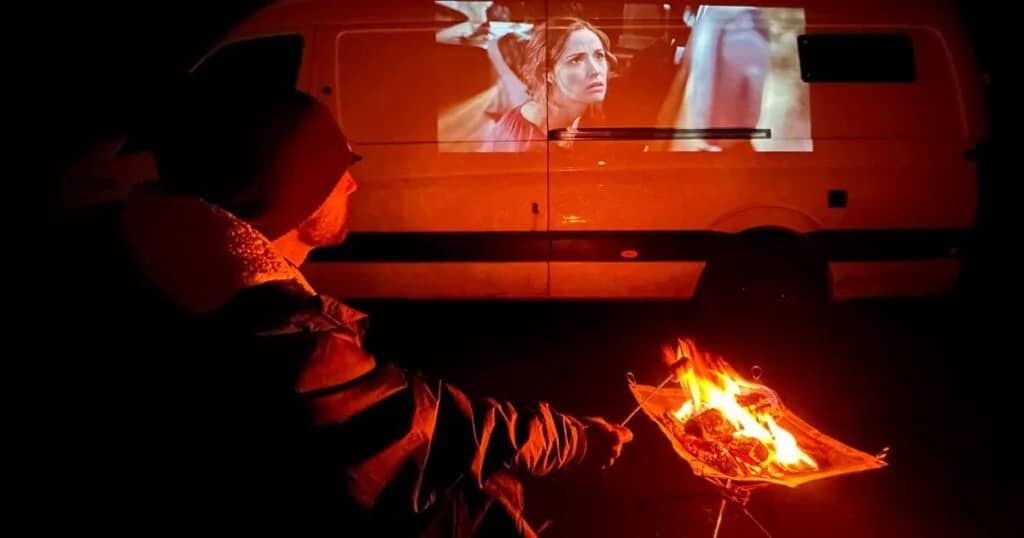 A man sat at a firepit, toasting marshmallows, while watching a horror movie projected onto the side of a campervan.