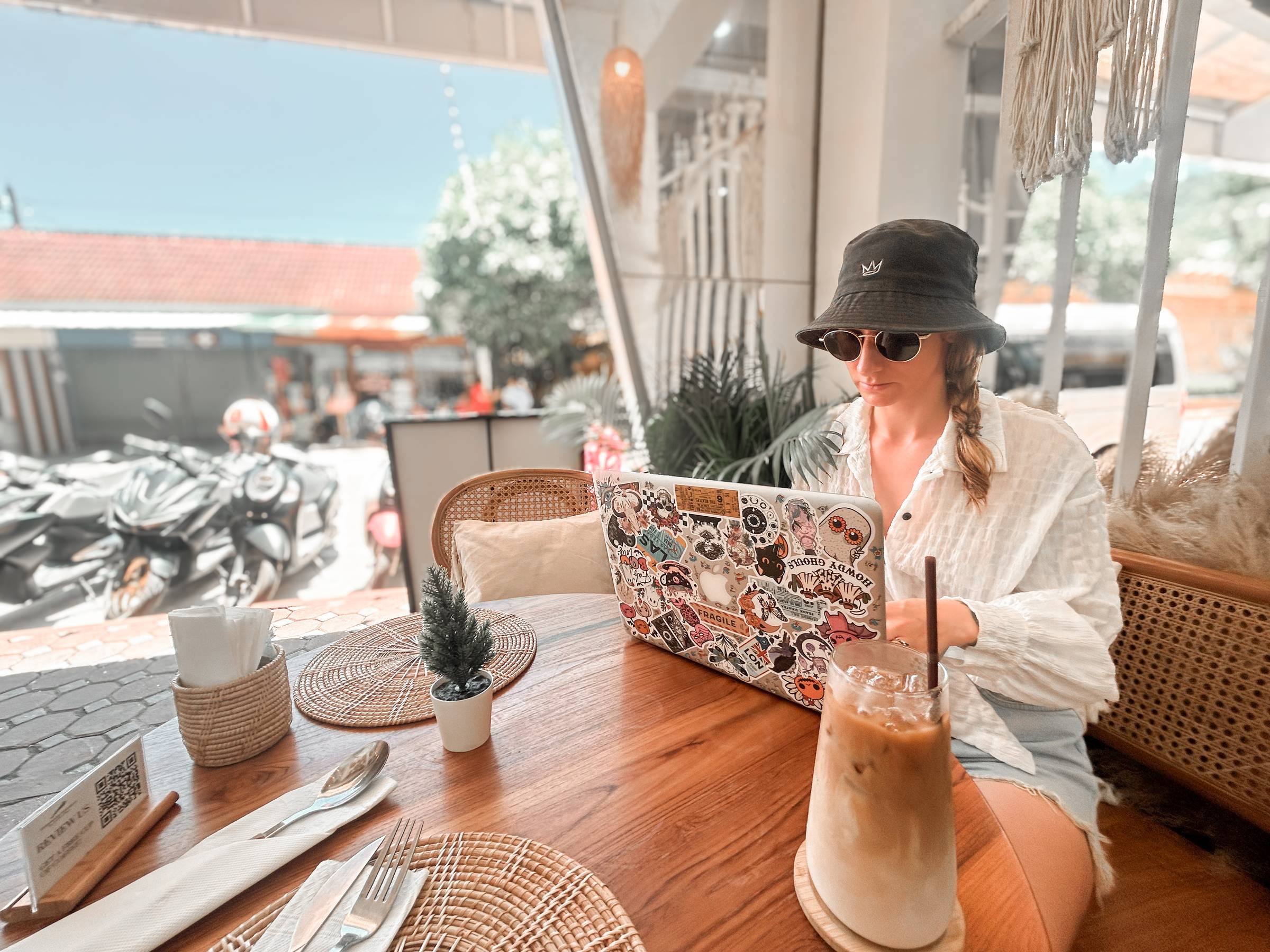 Woman sat at a laptop outside a cafe in Thailand with an iced coffee.