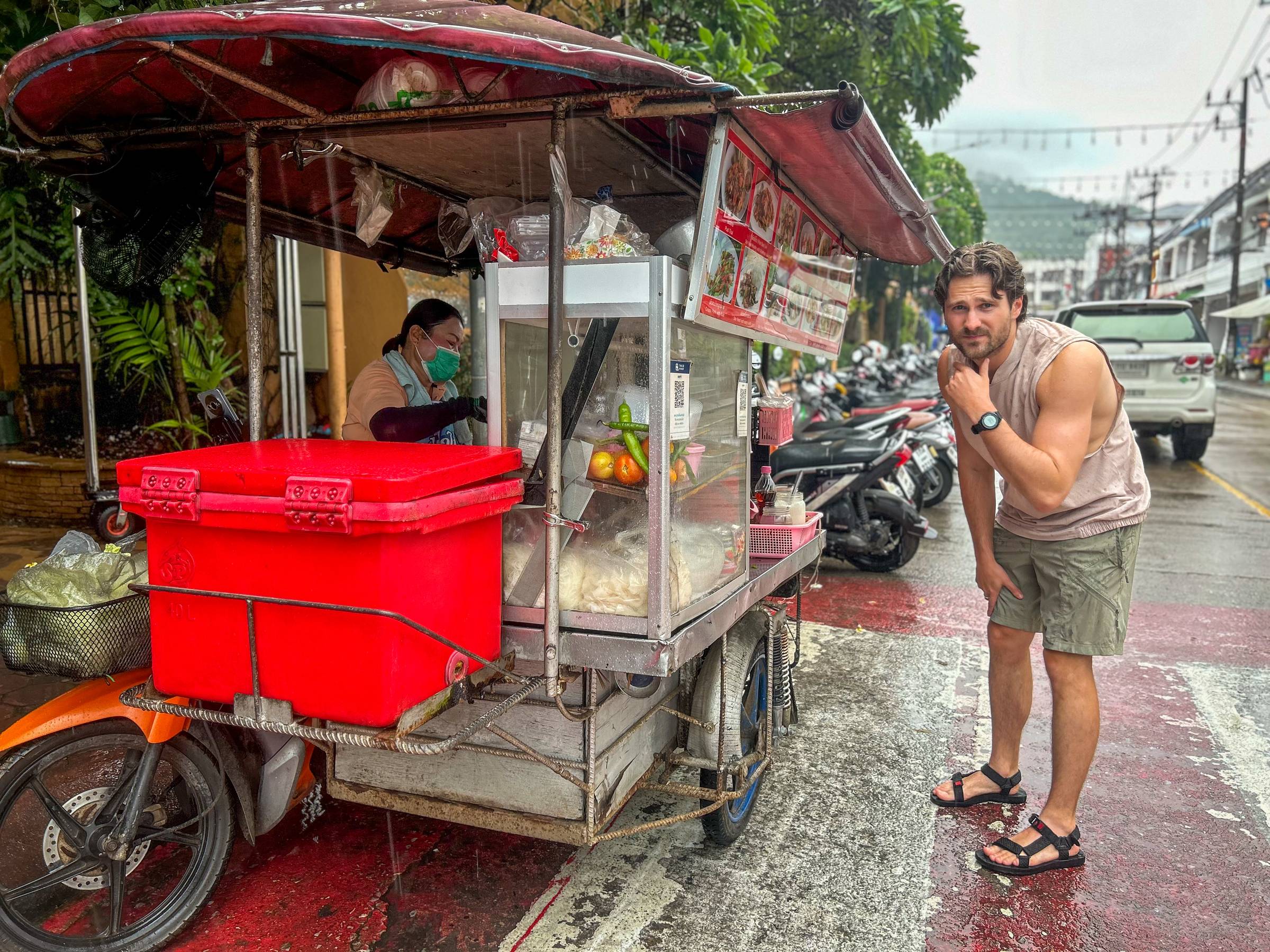 Man stood at scooter food cart in Phuket Thailand in the rain.