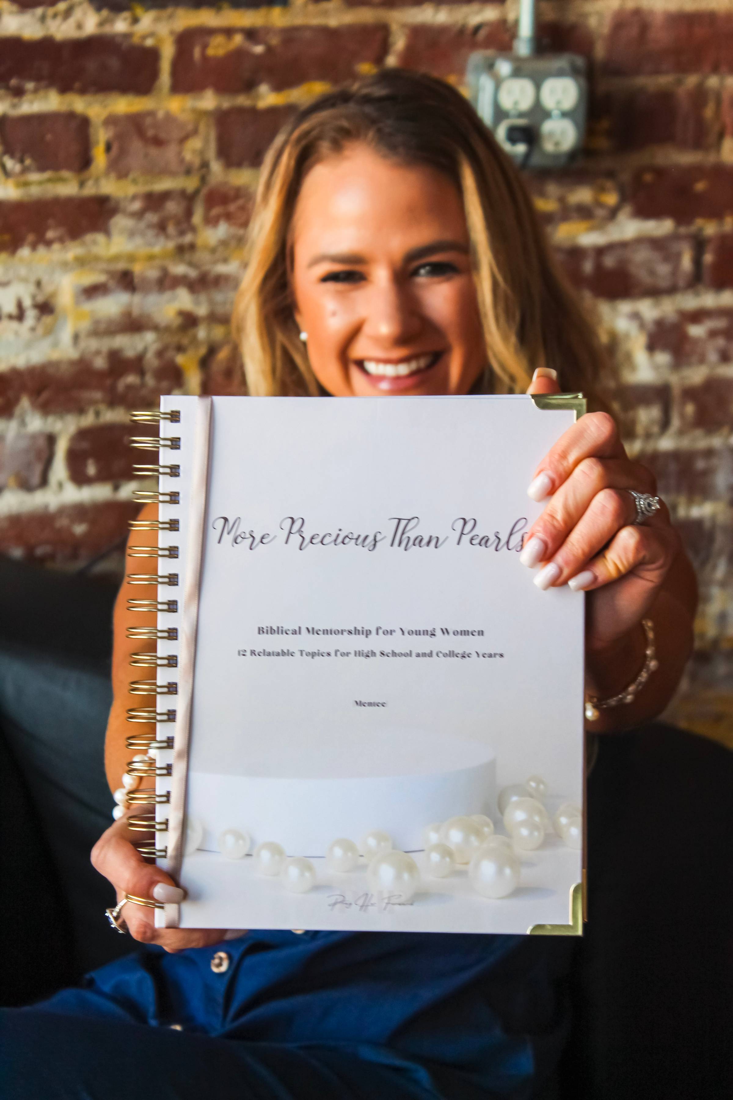 A person smiling and holding up a spiral-bound book titled "More Precious Than Pearls," subtitled "Biblical Mentorship for Young Women." The book cover features scattered pearls on a light background. The person is sitting in front of a brick wall.