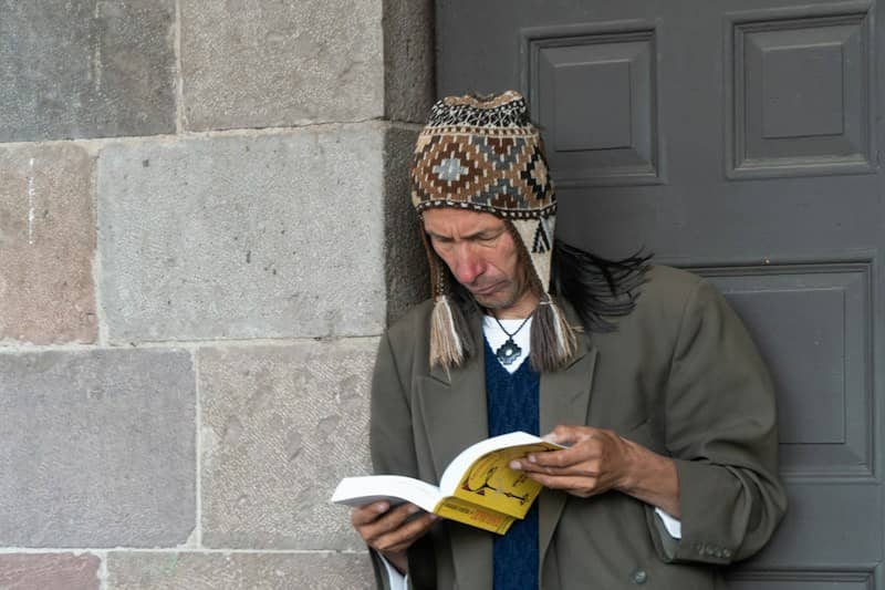 Hombre sentado leyendo un libro, con gorro tejido, frente a un muro de piedra.