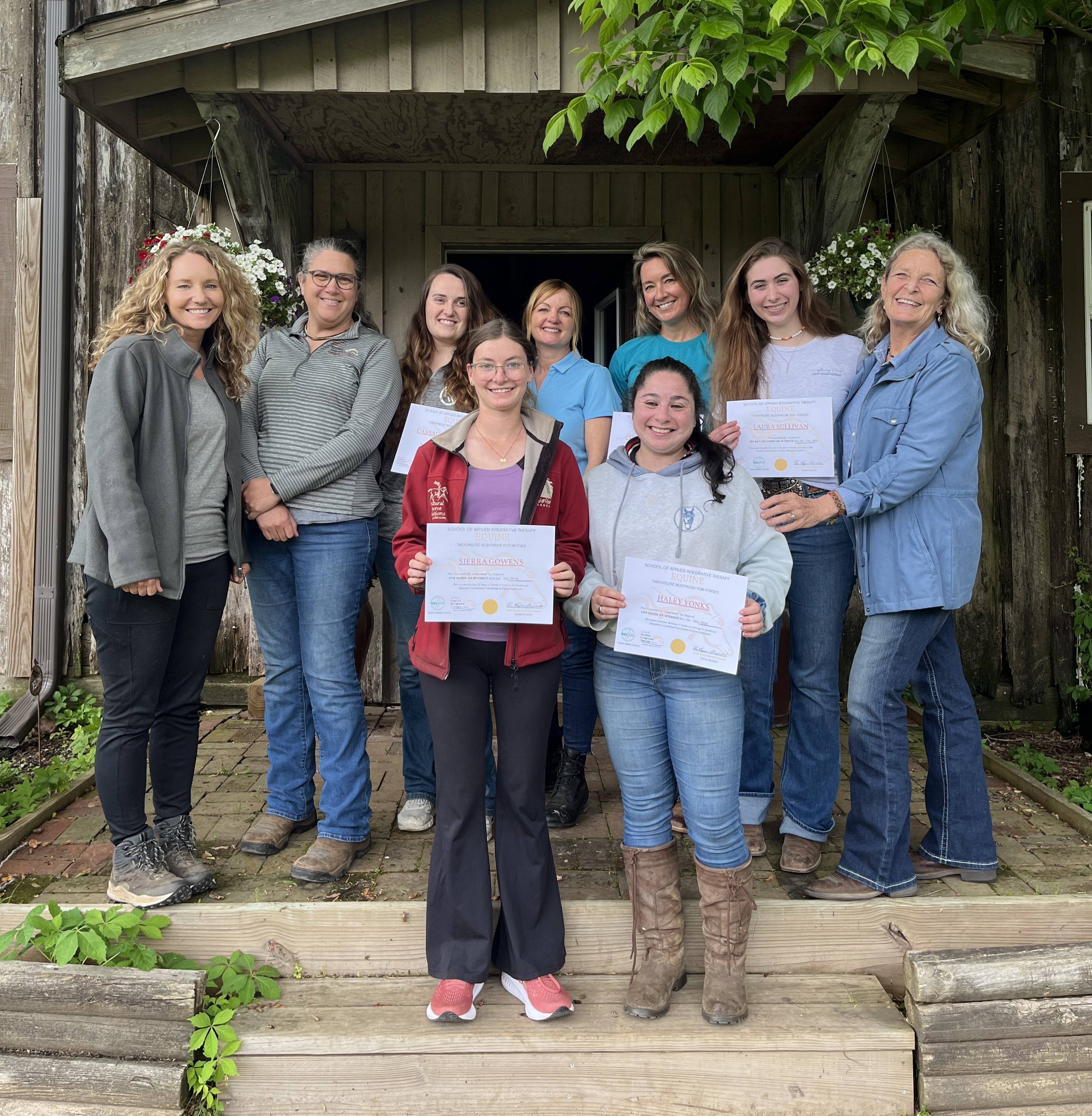 A group of eight people is standing on wooden steps in front of a rustic building. Three individuals in the front row are holding certificates. The setting is outdoors, with greenery and potted flowers visible. Everyone is smiling and dressed casually, suggesting a celebratory or graduation event.