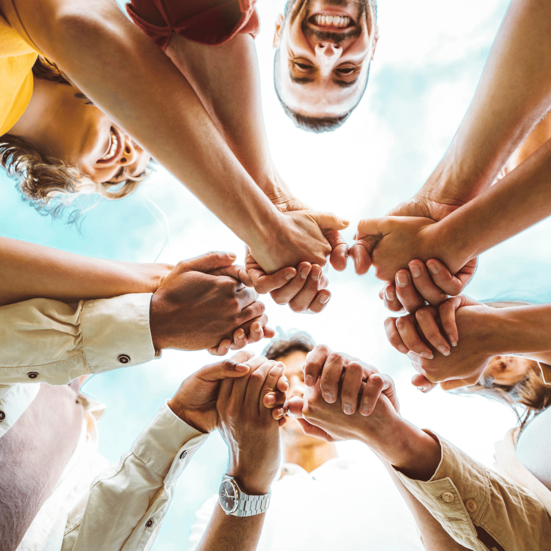 A diverse group of people standing in a circle, holding hands, and smiling, viewed from below against a bright sky, symbolizing unity and collaboration.