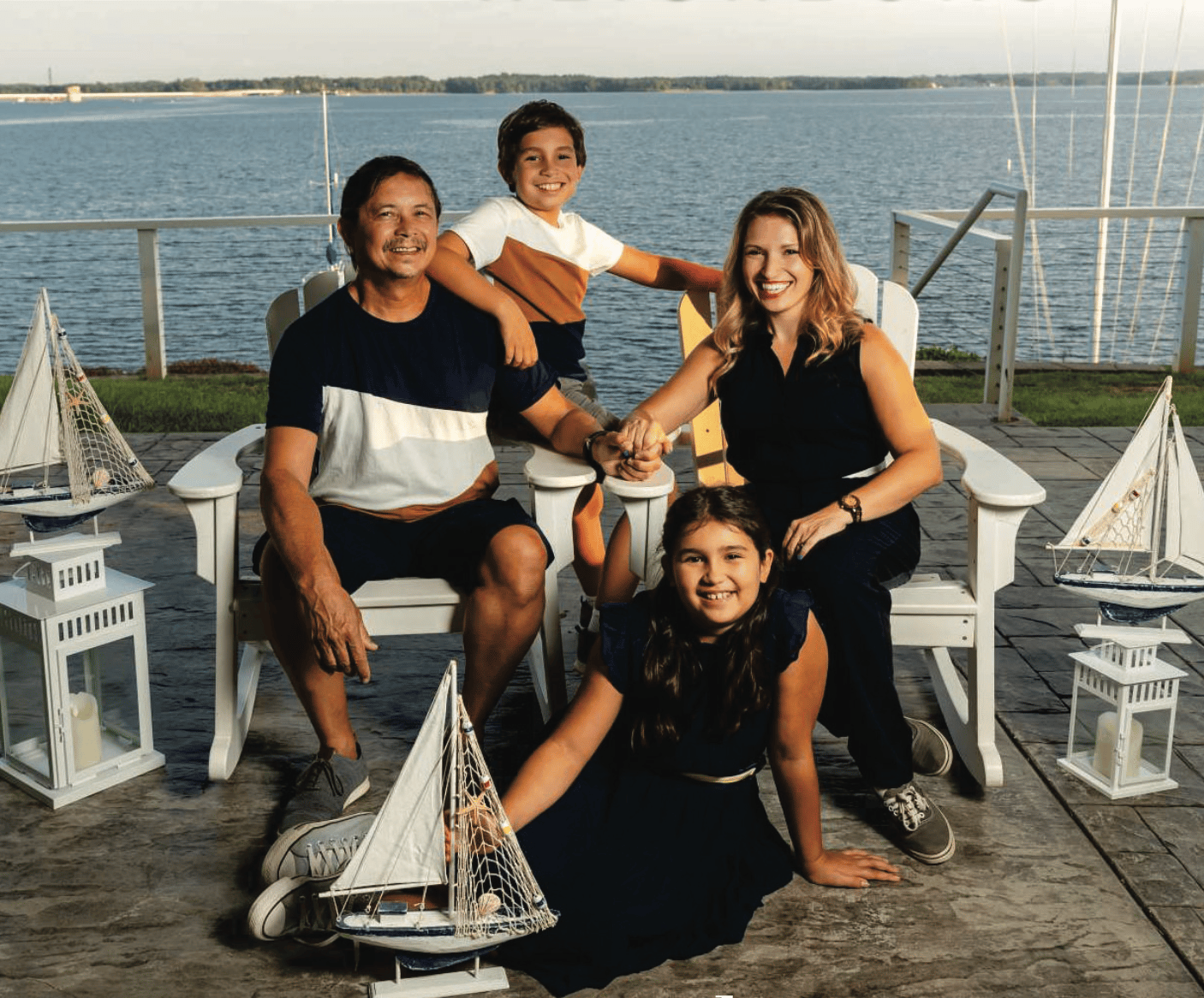 A smiling family of four seated outdoors near a waterfront, surrounded by nautical decor, including model sailboats and lanterns, on a sunny day.