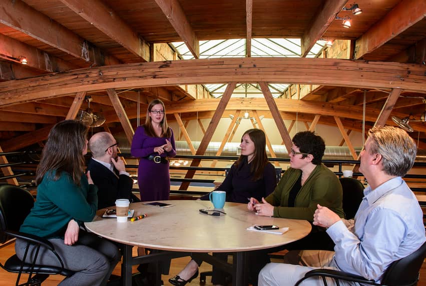 5 people in business casual attire sit around a round table, looking at a standing woman in a purple dress who appears to be delivering a presentation. In the background are skylights and wooden trusses from the interior of the building.