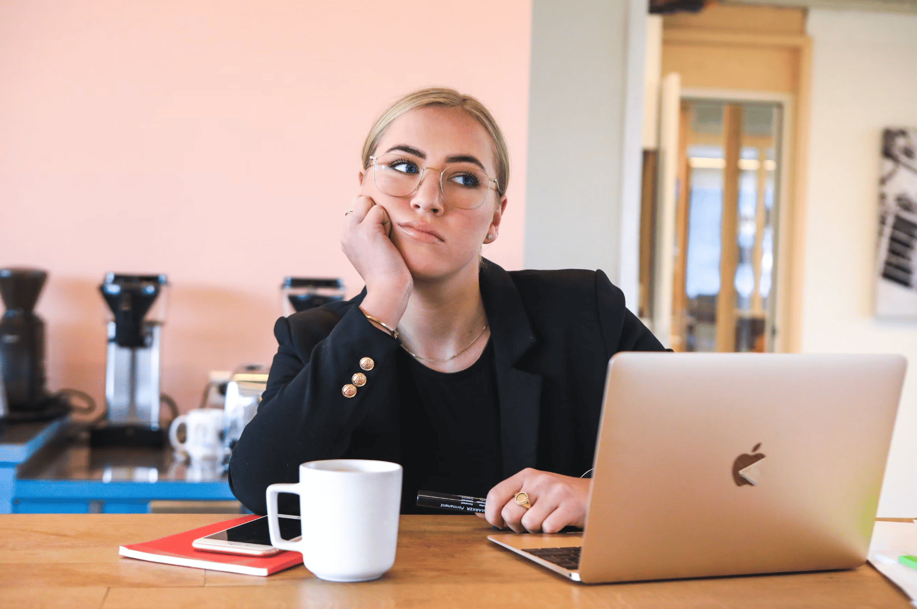 A white woman with blonde hair and glasses sits at a desk in front of macbook computer. She is resting her chin on her hand and has an expression of boredom in her eyes