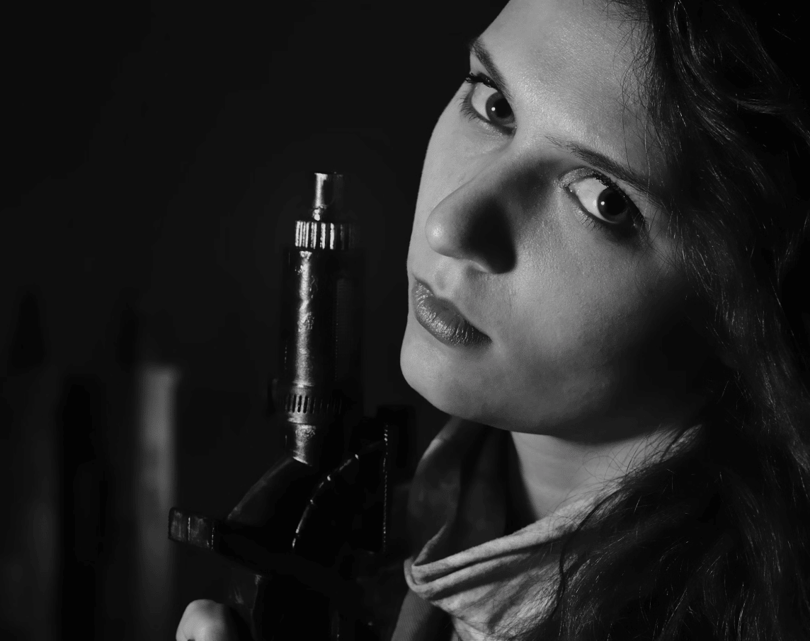 A black and white photo of a woman holding a gun pointing upwards. She is looking at the viewer with a sidelong glare.