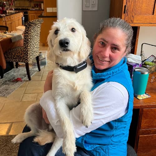 Fluffy white dog with a black collar sitting in the lap of a woman wearing a white shirt and a turquoise best. They are in the foreground of a kitchen.