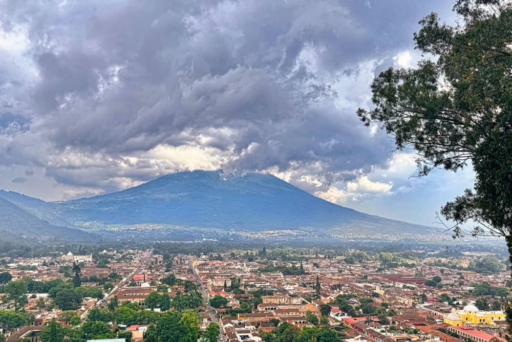 Hilltop view of Antigua, Guatemala and a volcano in the background. There are heavy dark rainclouds in the sky. 