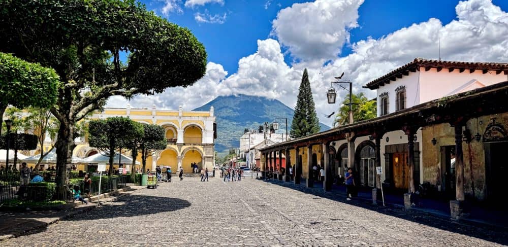 View from the public square in Antigua, Guatemala with bright yellow buildings and a cloud-covered volcano against a backdrop of blue sky. 