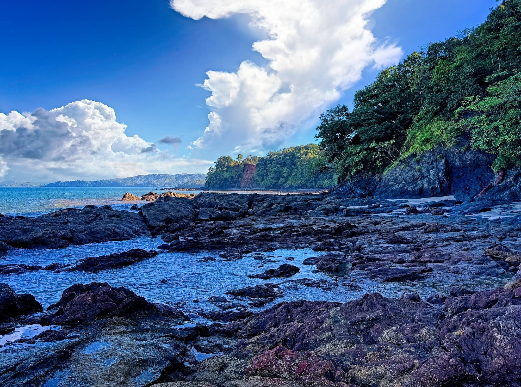 Remote beach with a rocky shoreline, clear water, dramatic clouds, and dense green coastline.