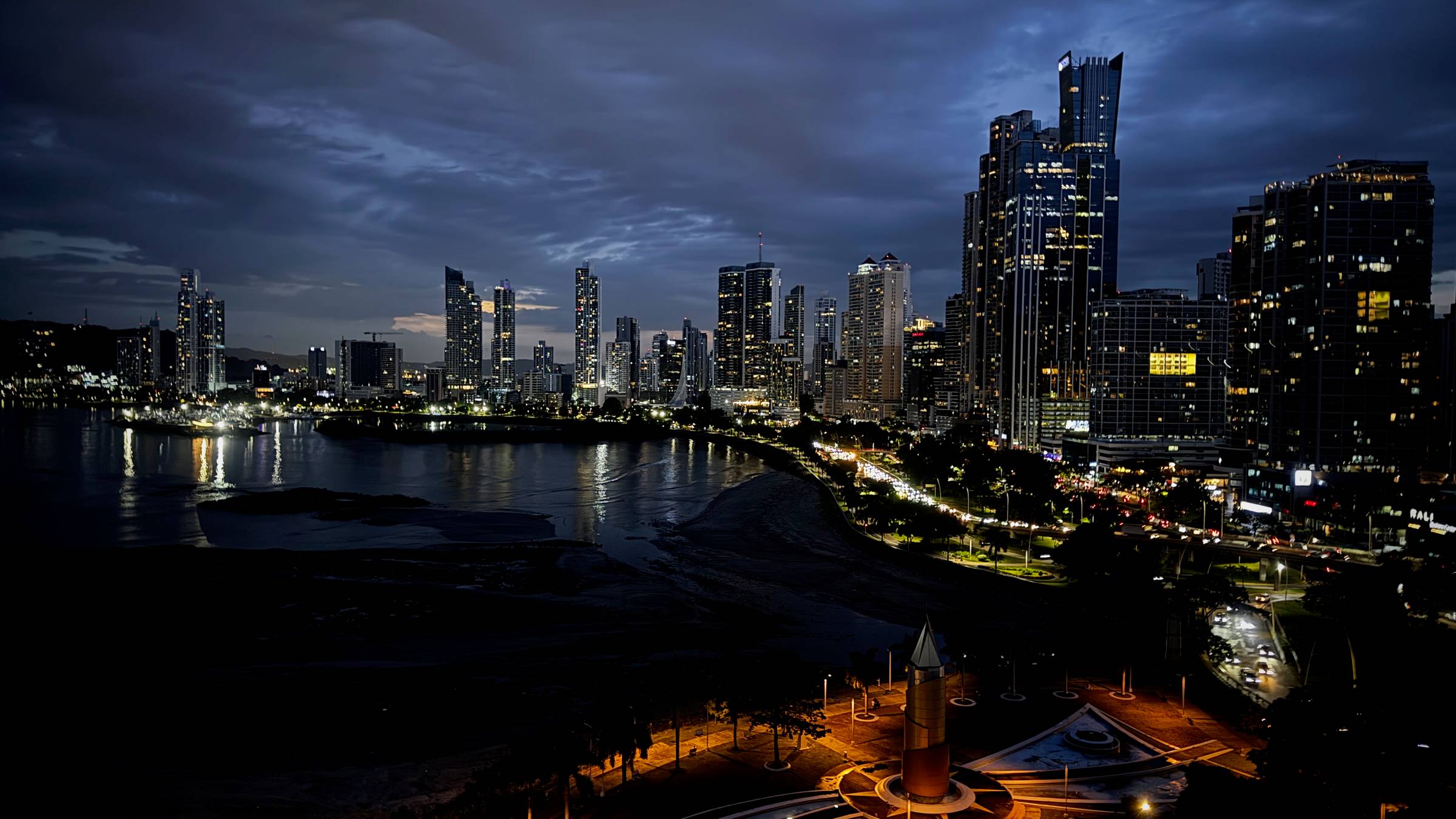 Skyline view of Panama City at dusk. Buildings are lit from within and lights reflect on the water of the ocean bay in the foreground. 