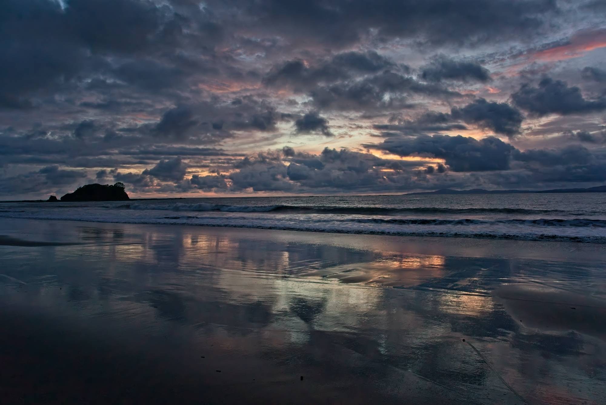 A cloudy beach sunset in hues of blue and pink. The wet shoreline reflects the colors. 