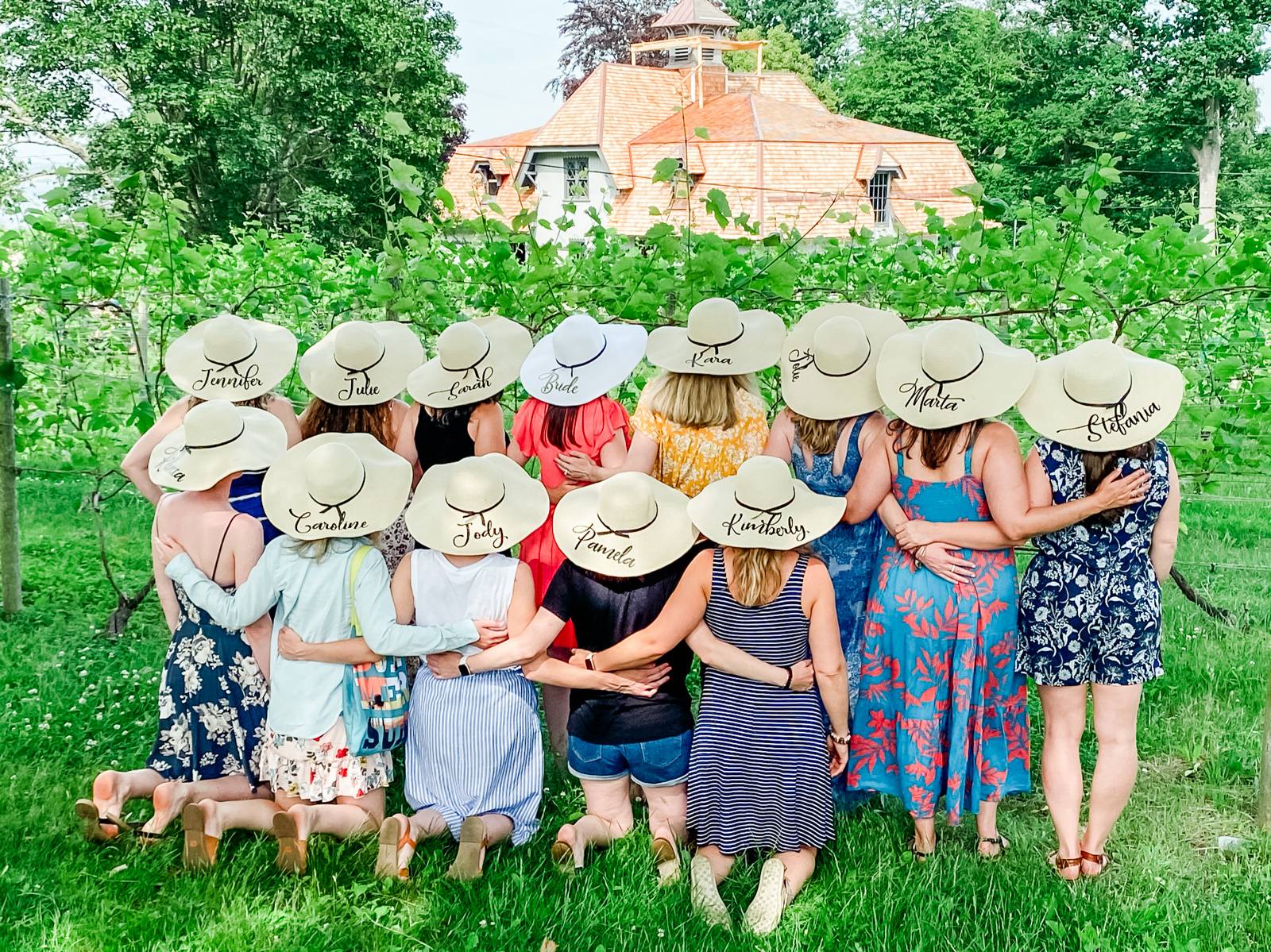 A group of women wearing sun hats embroidered with their names facing a green vineyard with a winery building in the background.