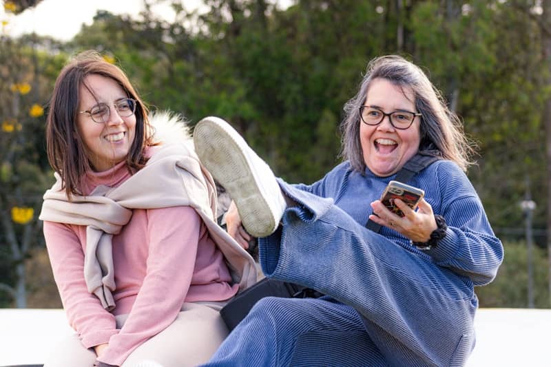 Two women laughing outdoors with one kicking their leg up.