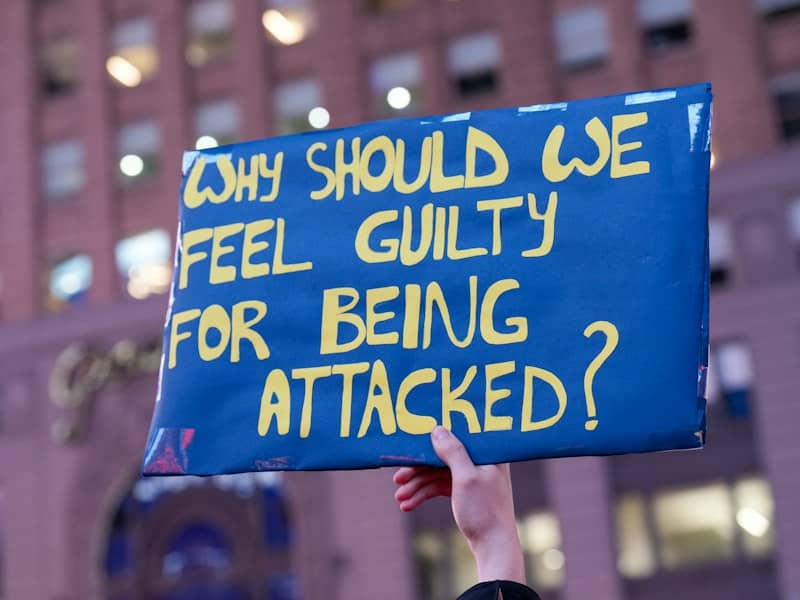 A hand holding a blue sign reading "WHY SHOULD WE FEEL GUILTY FOR BEING ATTACKED?"
