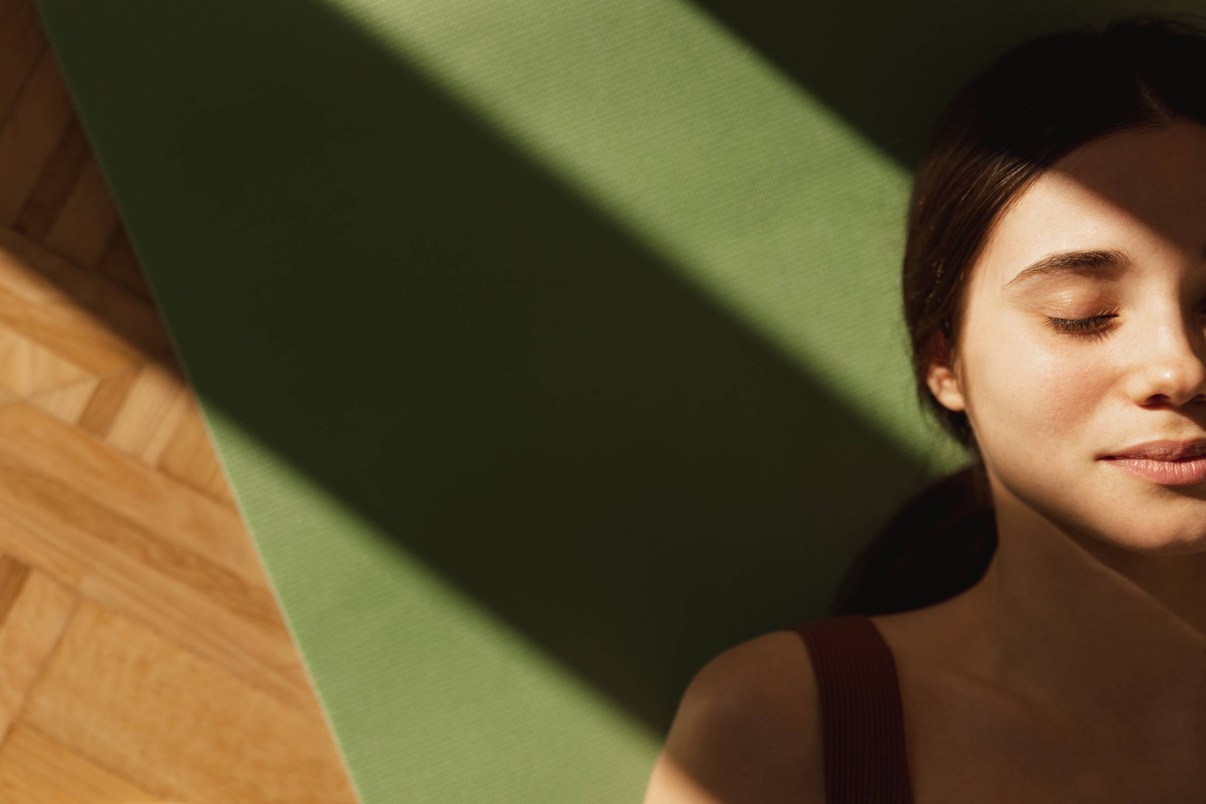 Une femme aux yeux clos dans un moment de sérénité, partiellement éclairée par un rayon de soleil. Elle se tient à côté d'un tapis de yoga vert sur un sol en bois.