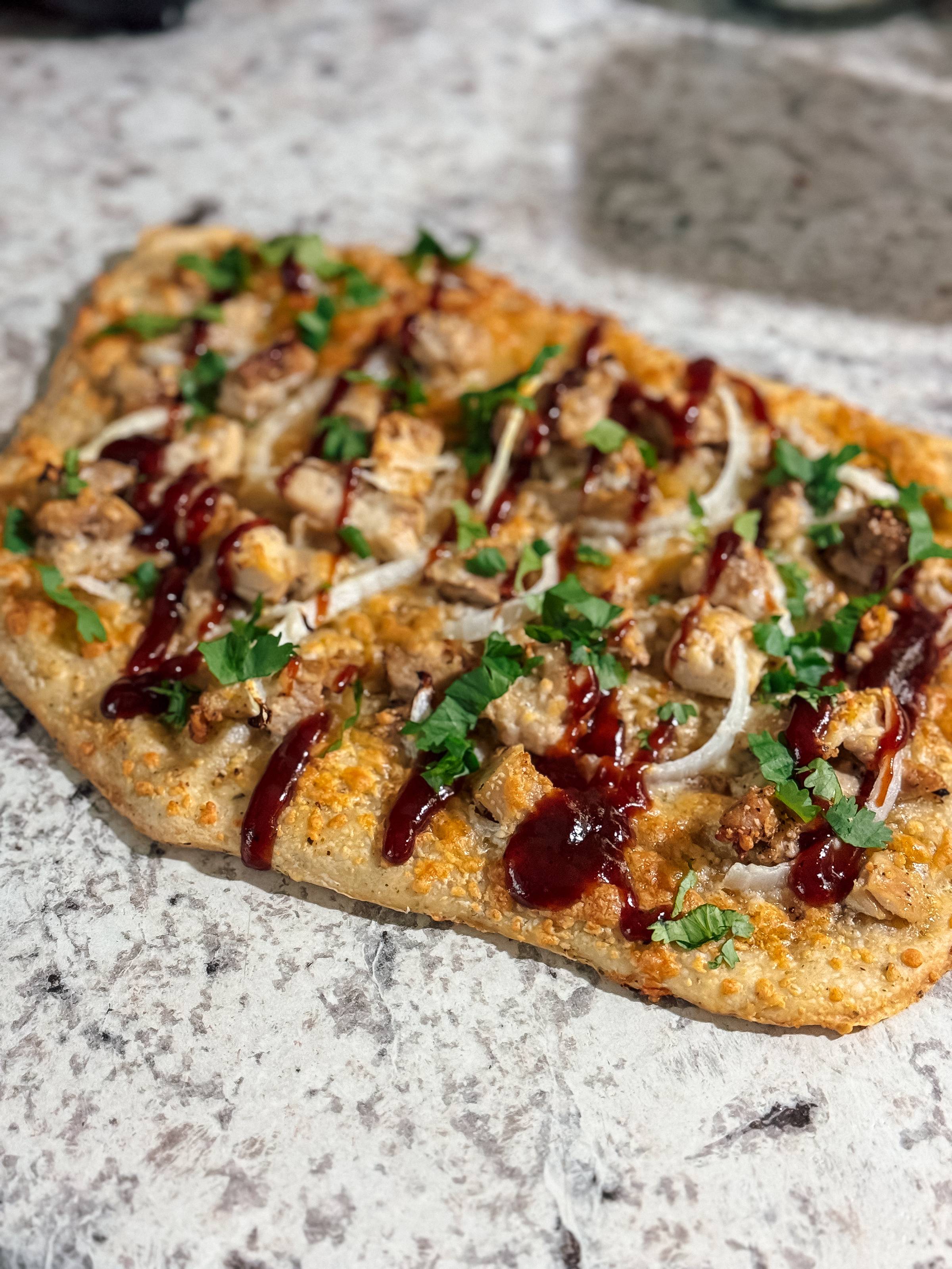homemade flatbread lies on a counter waiting to be cut. It is topped with chicken pieces, sticky sweet BBQ sauce, sliced onions, cheese & cilantro