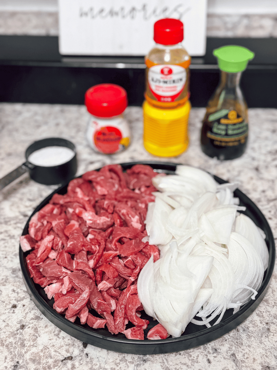   a large, circular black ceramic plate sits on a countertop. It is filled with sliced deep red uncooked meat cut into stips on the left half & thinly sliced white onions on the right half. Behind the plate is an array of other ingredients, one in a black