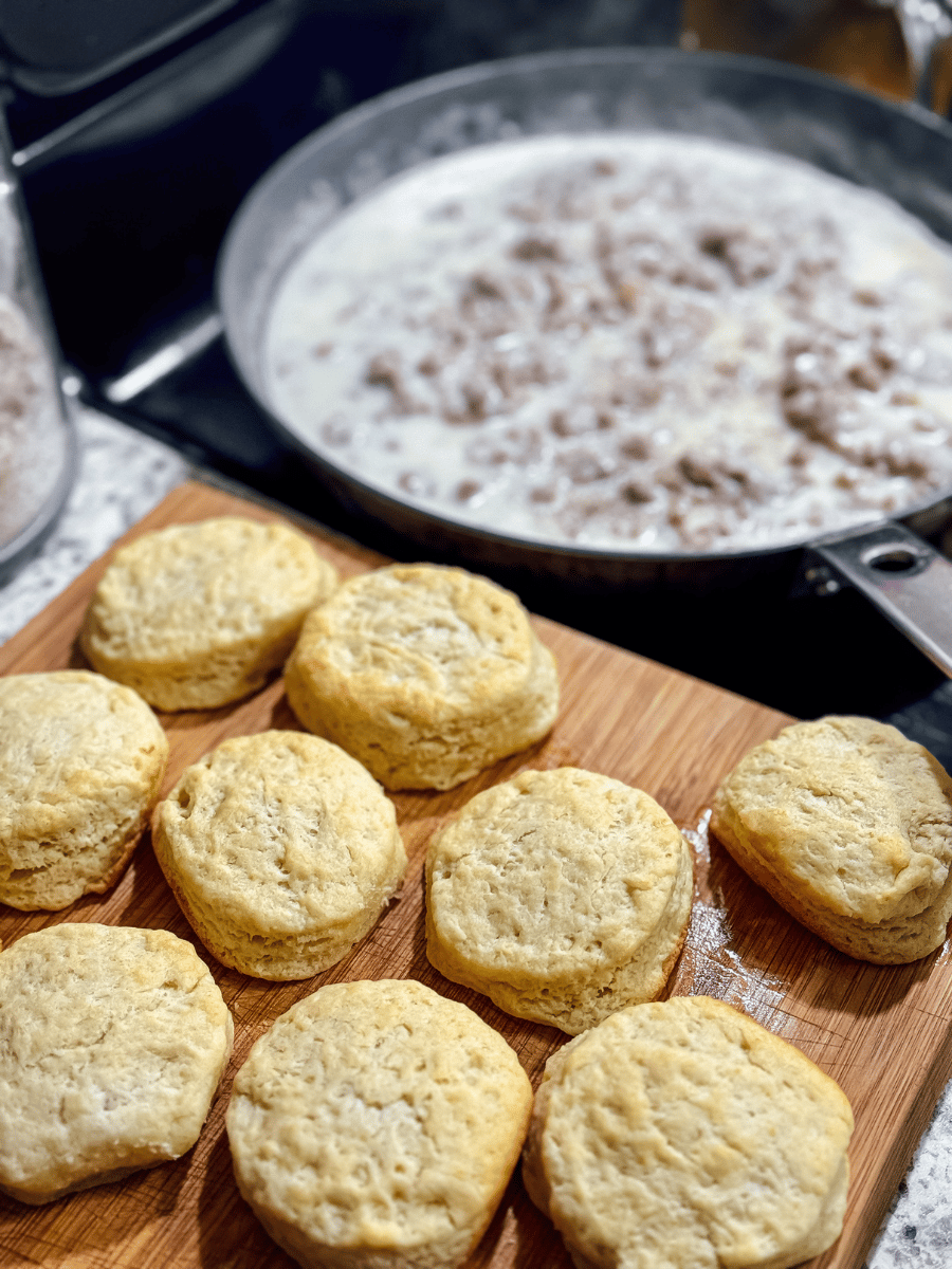 9 golden browned homemade biscuits sit on a wooden cutting board next to a stove. There is a large skillet of sausage gravy sitting on the stove.
