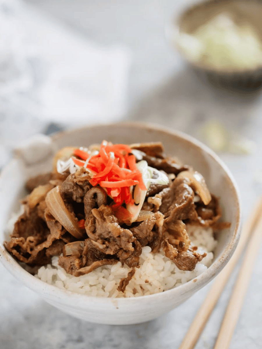 a small white bowl sits on a countertop filled with white rice that's topped with browned beef slices & onions & pickled pink ginger. Wooden chopsticks lie beside the bowl.