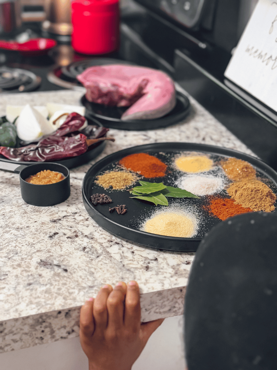 A small child stands looking up onto a counter, likely from tiptoes as their hands are holding on to the edge of the counter. A large ceramic circular black plate lies on the countertop & is filled with tiny piles of colorful spice powders, bay leaves, cloves & black peppercorns. A smaller circular ceramic black plate lies next to it holding onions, green jalapeno peppers & red chile peppers. A small measuring cup of minced garlic lies between them. On a third small circular ceramic black plate lies an uncooked, pink cow tongue.