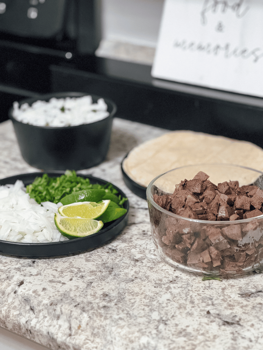 A self-serve style taco spread is laid on a kitchen countertop. There is a small circular black ceramic plate with diced white onions, bright green cilantro & sliced lime wedges. A transparent glass bowl filled with browned cubed meat. A stack of tortilla