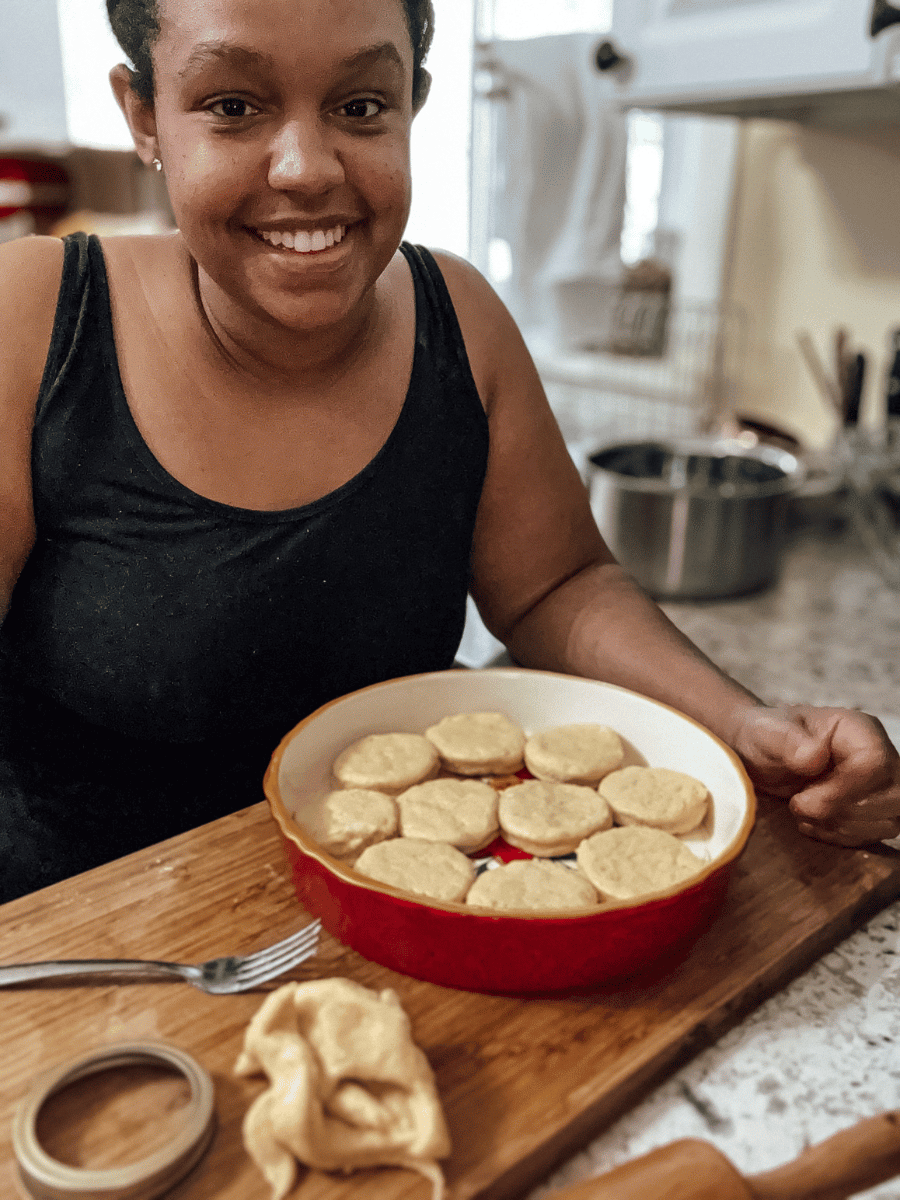 brown girl in a blank tank top stands besides a wooden cutting board that has a red pie dish filled with circular biscuits ready for baking. There is scrap biscuit dough, a canning lid ring, fork lying on the same cutting board as evidence of making homemade biscuits. A wooden rolling pin can also be seen resting on the counter beside the cutting board.
