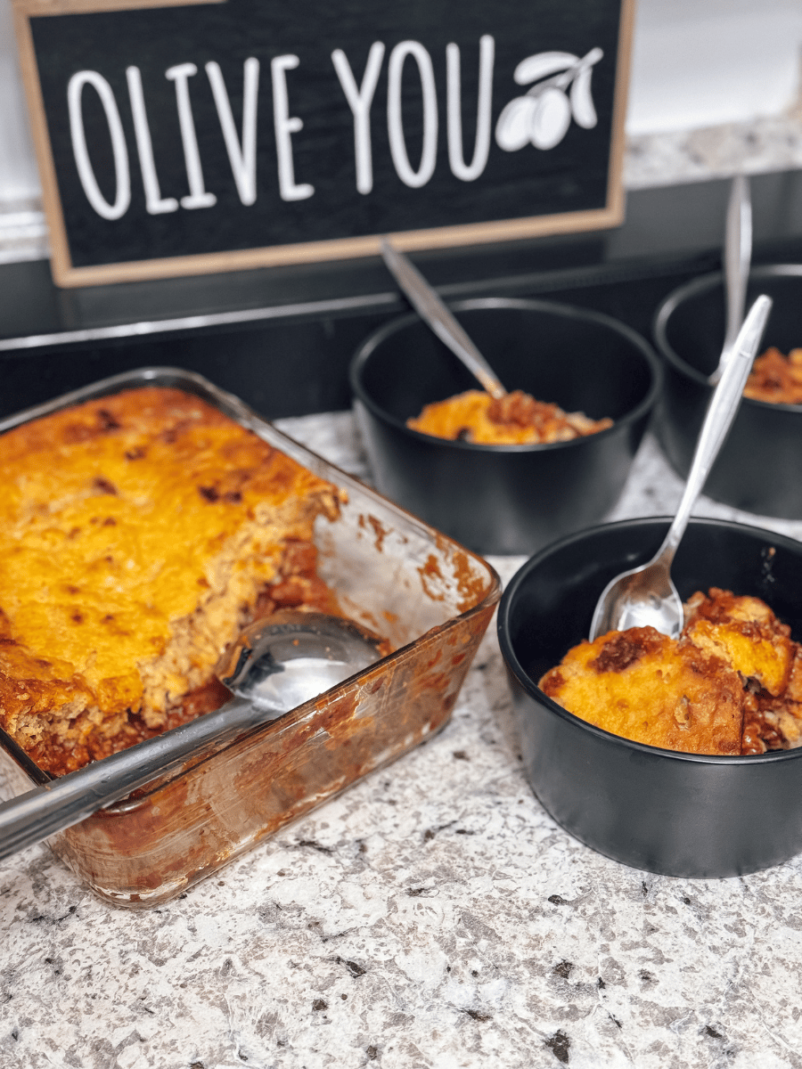 a transparent glass casserole dish is 3/4 full of a comforting meal of tamale pie. Red chili on the bottom is topped with golden yellow cornbread. There is a large stainless steel ladle in the casserole dish for scooping & 3 small black ceramic bowls with servings of the tamale pie beside the casserole pan.