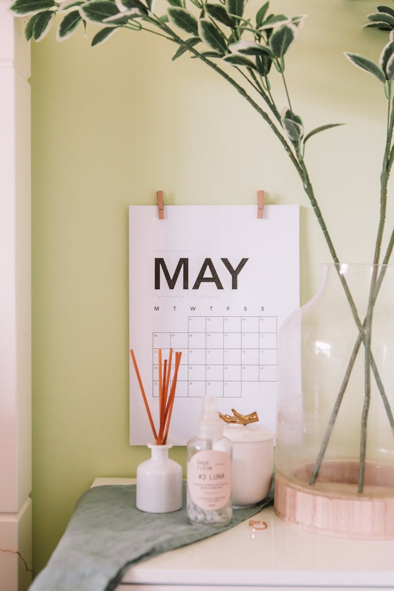 "A calendar showing the month of May is pinned against a yellow wall above a desk, which is adorned with a diffuser, simple flowers, and other items, creating a serene workspace setting."