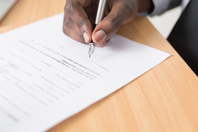 "Close-up of a hand holding a pen, completing paperwork on a wooden desk, symbolizing employee onboarding or administrative tasks."