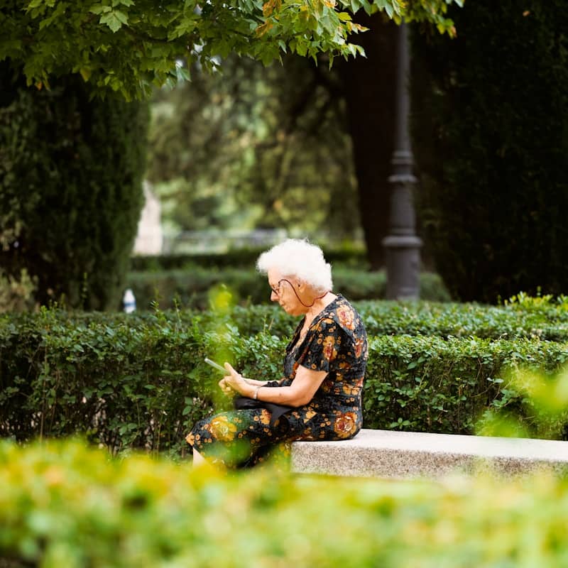 An elderly woman in a floral dress using a smartphone on a stone bench