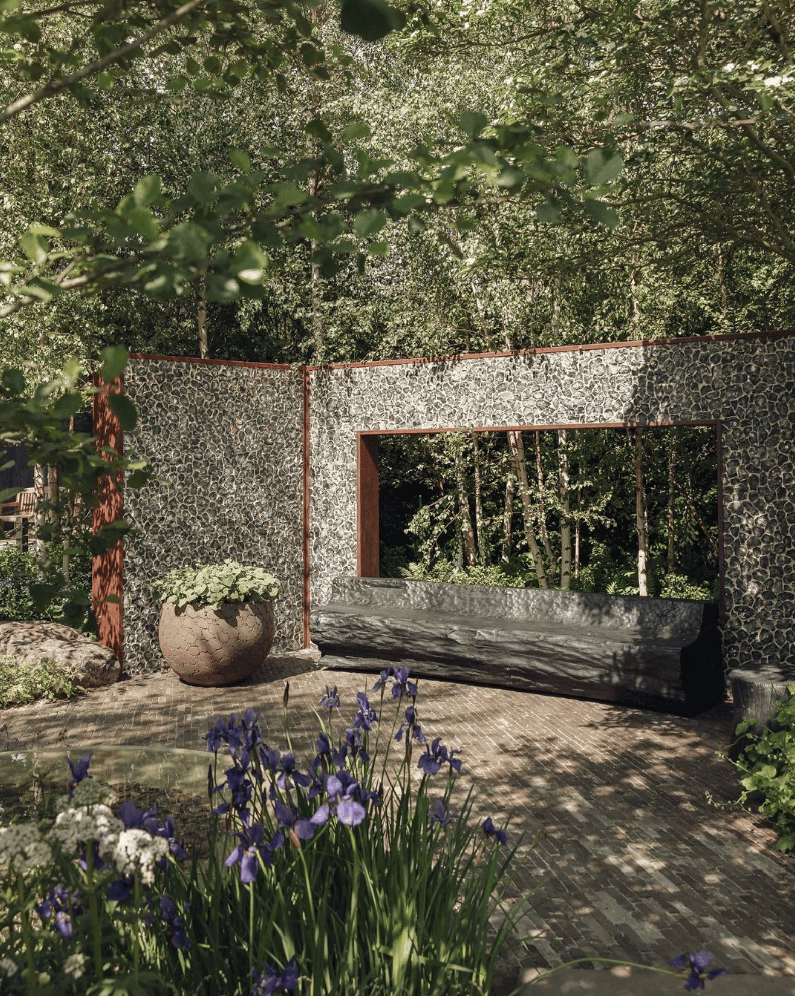 Ula Maria's Shinrin-Yoku Garden. There is a beautiful stone wall behind a hewn stone bench, surrounded by birch trees. A large, rounded brown pot full of plants sits next to the bench.