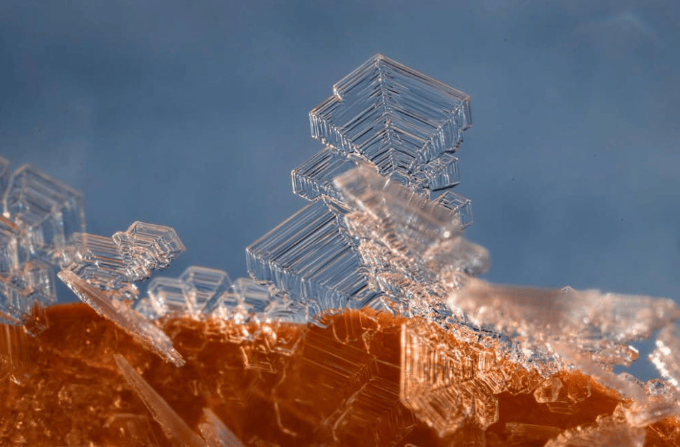 A macro shot of ice crystals forming on a red rock with blue sky behind it.