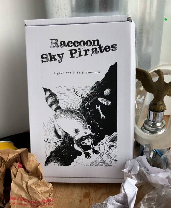 White mailer box situated amidst trash, with a drawing of a raccoon on it and the words Raccoon Sky Pirates