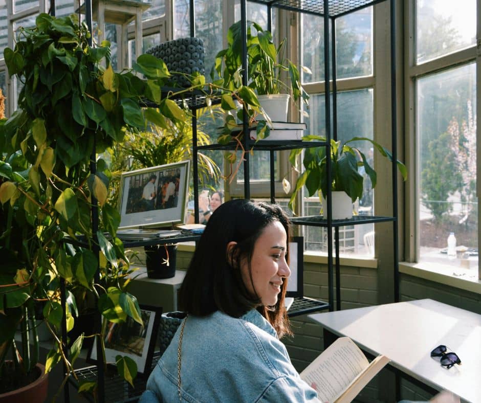 A woman reading at a desk in front of a window surrounded by plants.