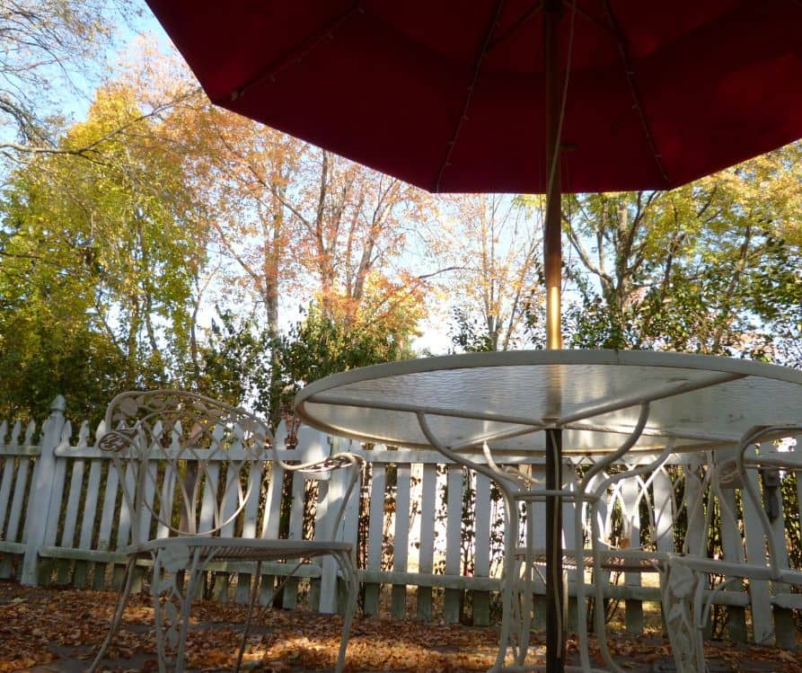 Fall backyard sitting area with white metal chairs, a table and red umbrella next to a white picket fence.