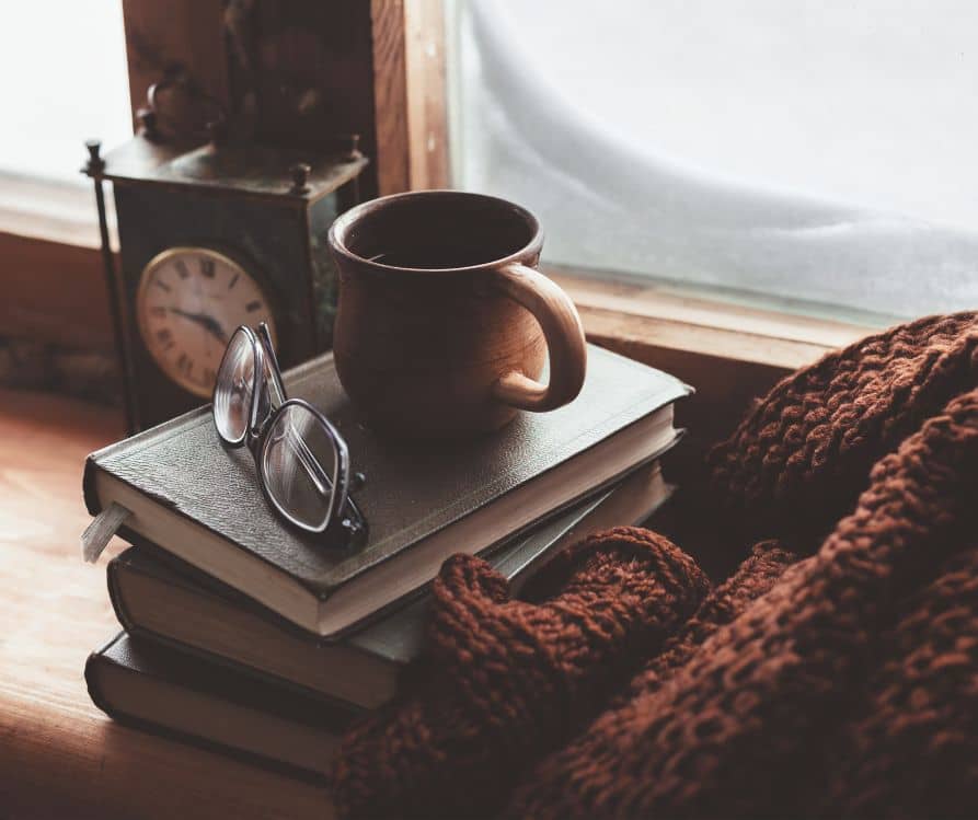 A cozy brown blanket, a stack of books, a natural mug and glassed next to an insulated winter.