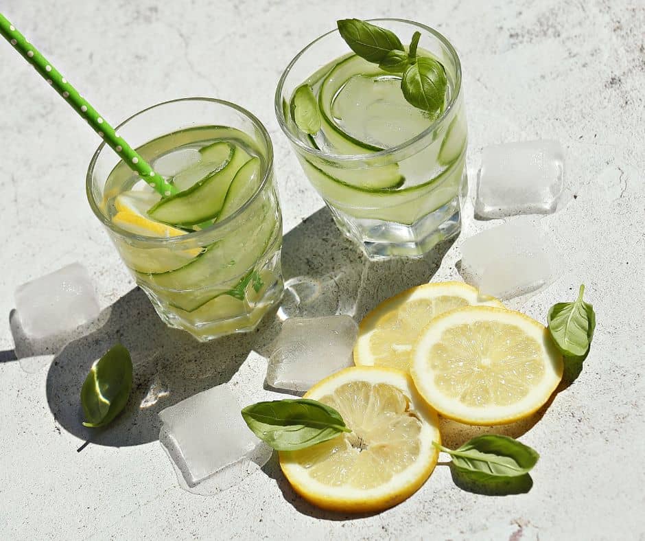 Basil cumber coolers with lemon in clear glasses on a grey countertop with lemon slices and ice cubes.