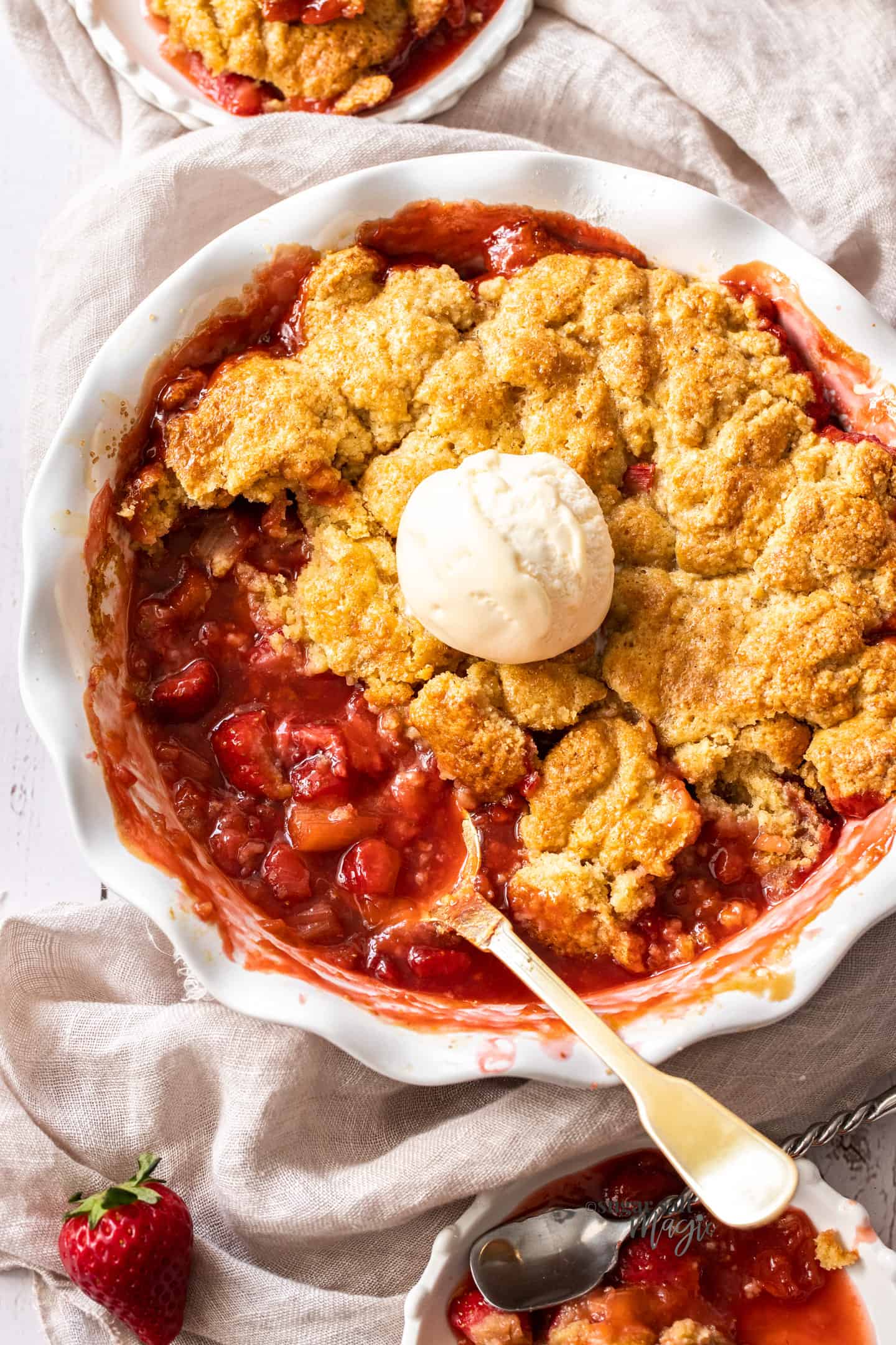 Top down view of a strawberry rhubarb cobbler with a spoon sticking out of the dish and some of the topping missing.
