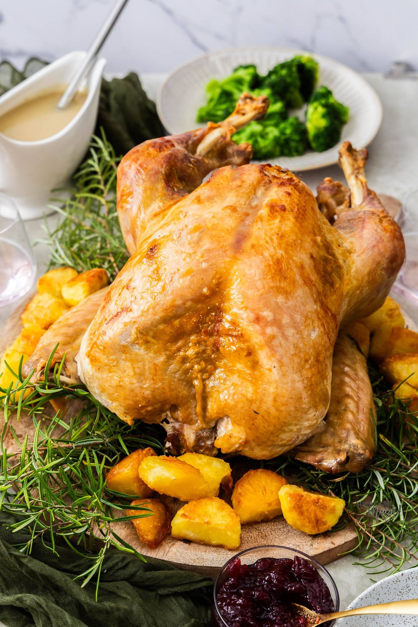 A whole roast turkey on a wooden board with potatoes and rosemary.