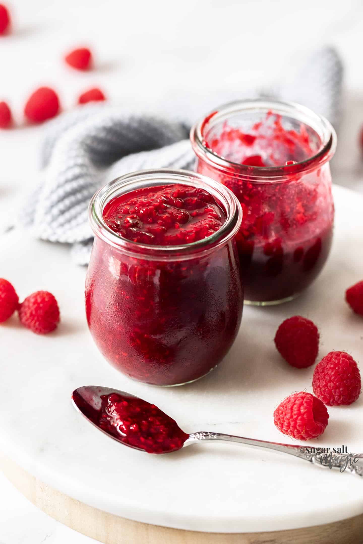 Two glass jars of raspberry compote on a marble platter surrounded by fresh raspberries.