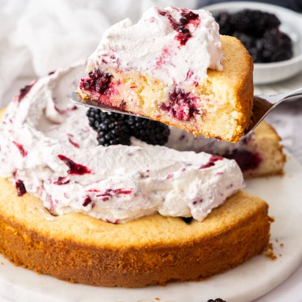 A slice of lemon cake dotted with blackberries and topped with cream being lifted from a full cake.