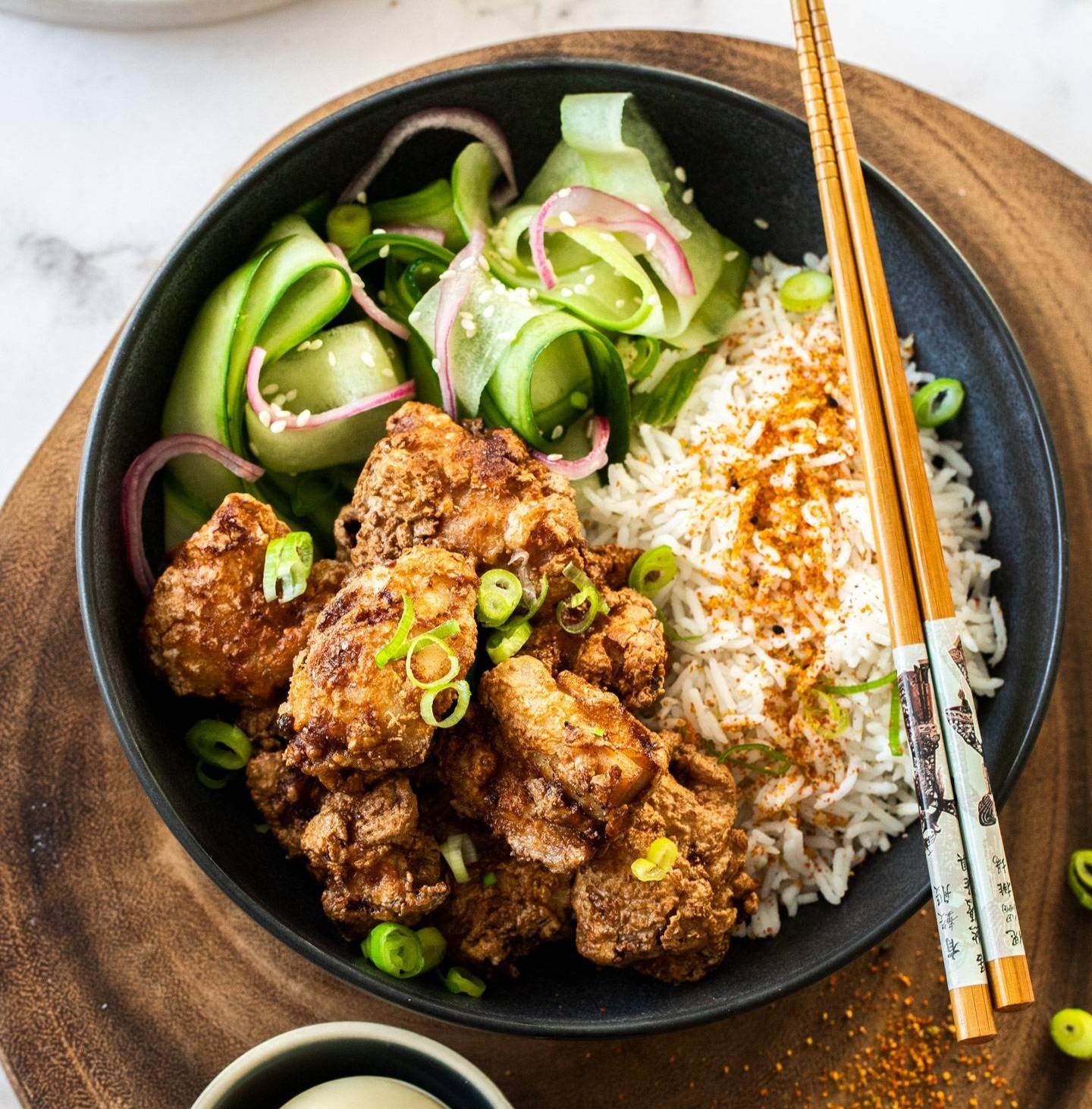 Top down view of a bowl with fried chicken, rice and cucumber with chopsticks across the top.