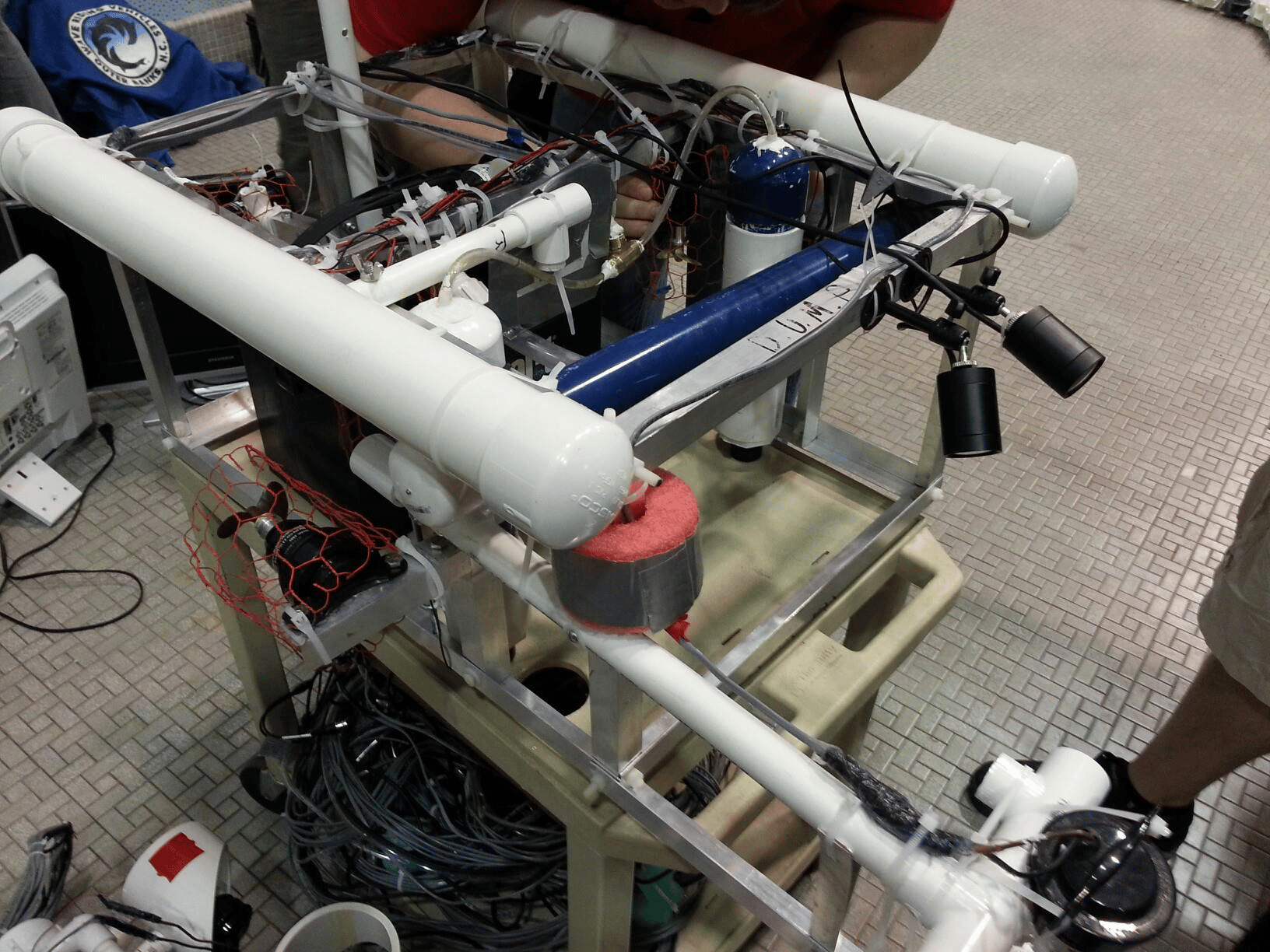 A complex robotics project built from PVC pipes, aluminum framing, wires, and mechanical components, assembled on a wheeled cart in robotics class.