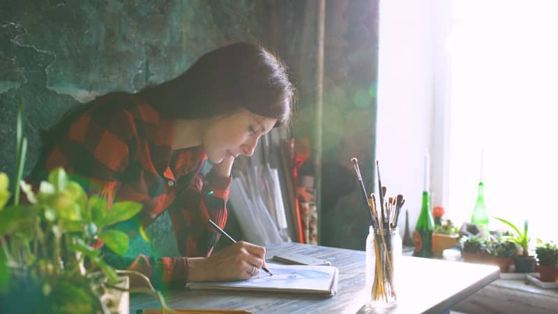 Woman painting at a desk with plants and sunlight.