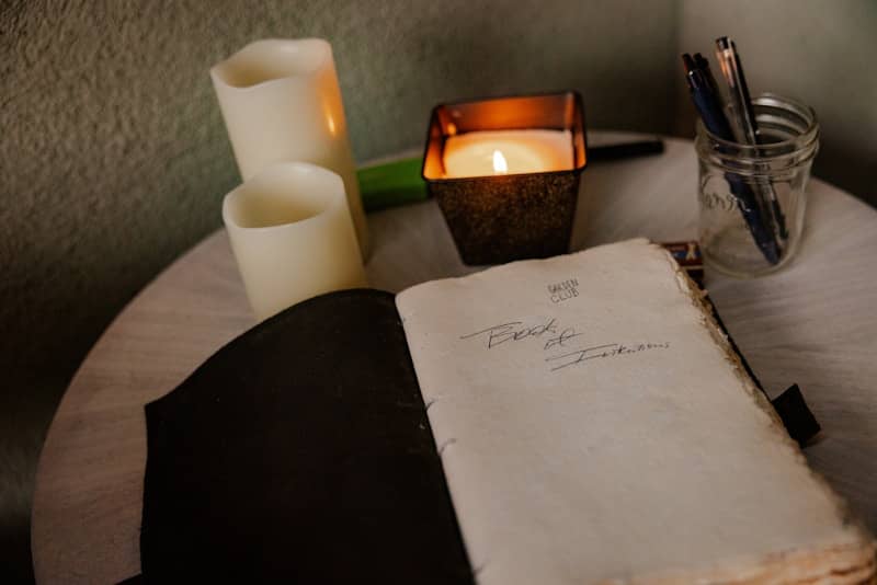 An open journal with handwritten text beside candles and pens in a glass jar